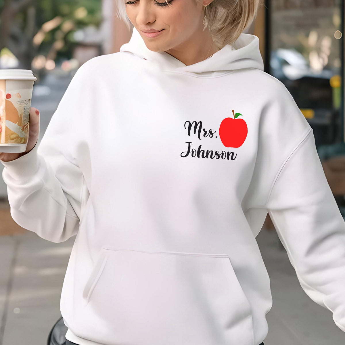 May include: White hooded sweatshirt with a front pocket. The sweatshirt features the text "Mrs. Johnson" in black script with a red apple graphic. A coffee cup is held in the left hand.