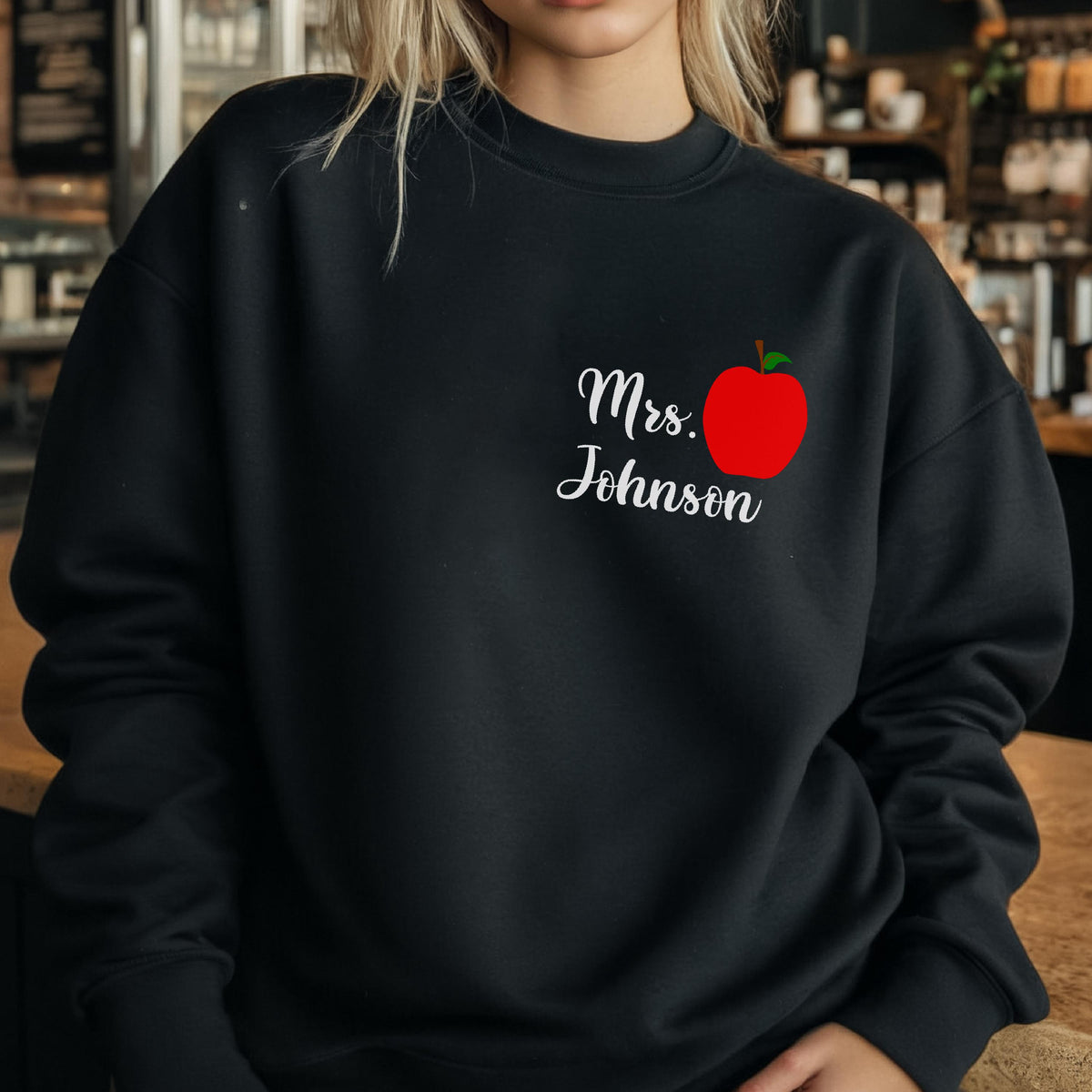 May include: Black sweatshirt with the text "Mrs. Johnson" in white script and a red apple graphic. The apple has a green stem. The sweatshirt is a crew neck style.