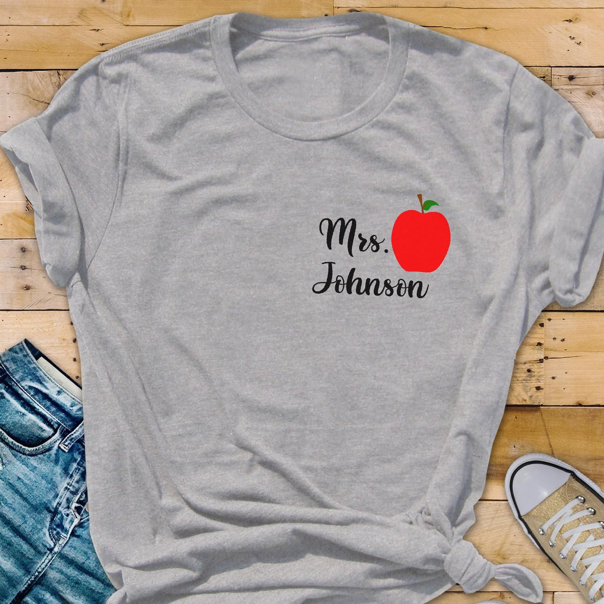 May include: A light gray t-shirt featuring the text "Mrs. Johnson" in black script, accompanied by a red apple graphic. The shirt is a casual style, with rolled sleeves and a knotted hem. The design is likely for a teacher.