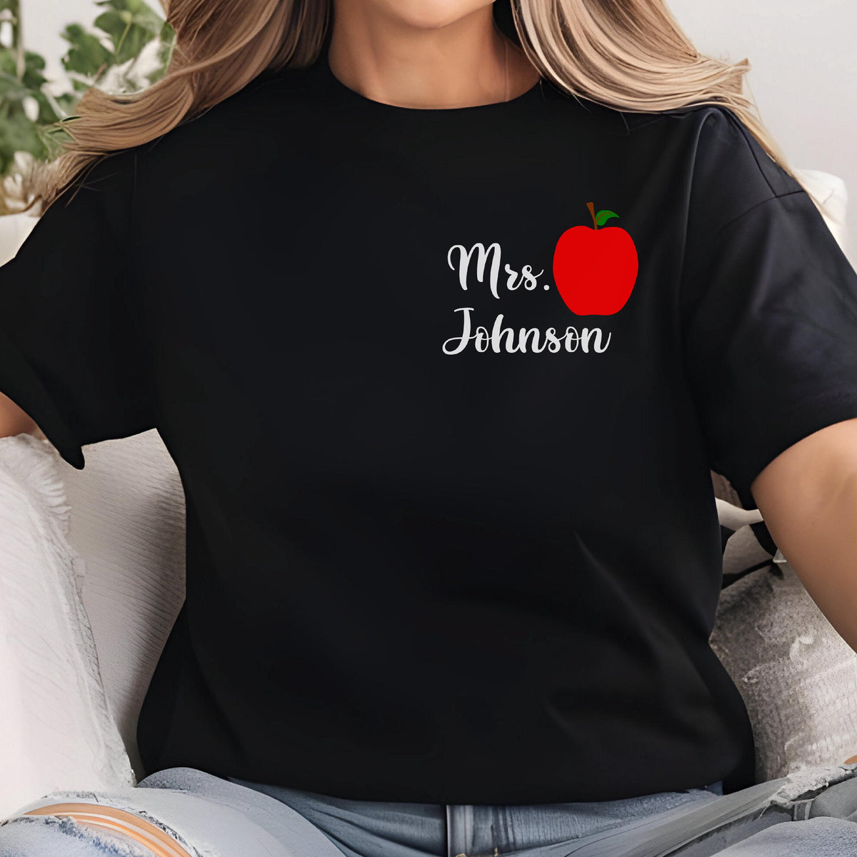 May include: Black t-shirt featuring a red apple graphic with a green stem and the text "Mrs. Johnson" in white script. The design is on the left chest area of the shirt. The shirt is made of a soft material.