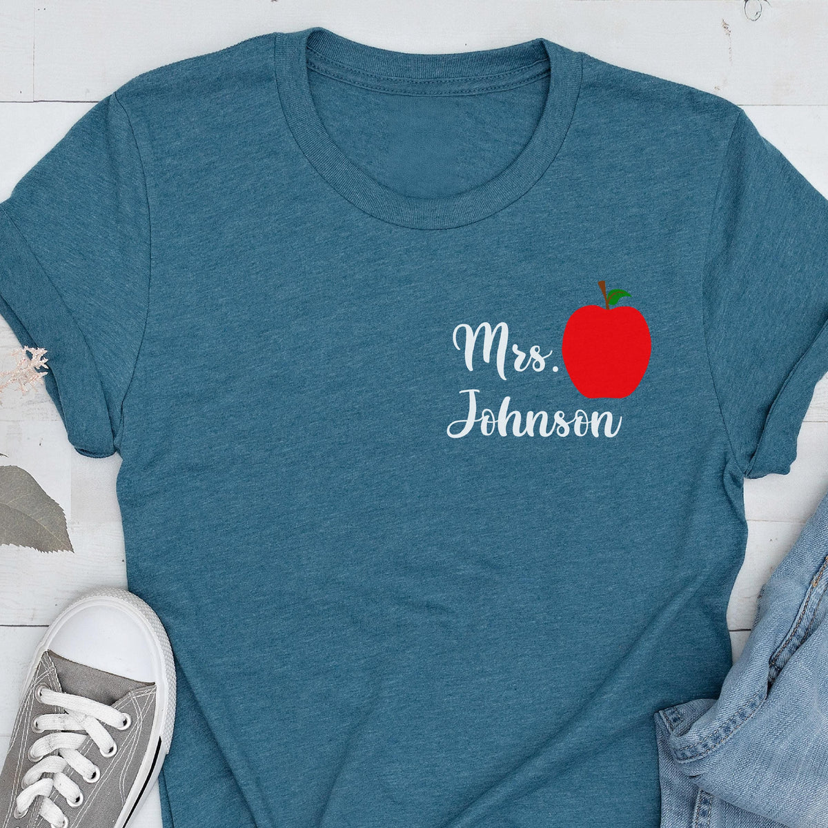 May include: Teal t-shirt with the text "Mrs. Johnson" in white script and a red apple graphic. The apple has a green stem and leaf. The shirt is made of a soft material.