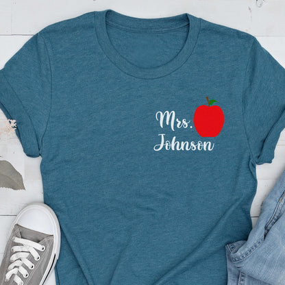 May include: Teal t-shirt with the text "Mrs. Johnson" in white script and a red apple graphic. The apple has a green stem and leaf. The shirt is made of a soft material.