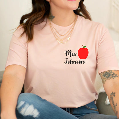 May include: A light pink t-shirt featuring the text "Mrs. Johnson" in black script, accompanied by a red apple graphic with a green stem and leaf. The shirt is a crew neck style.