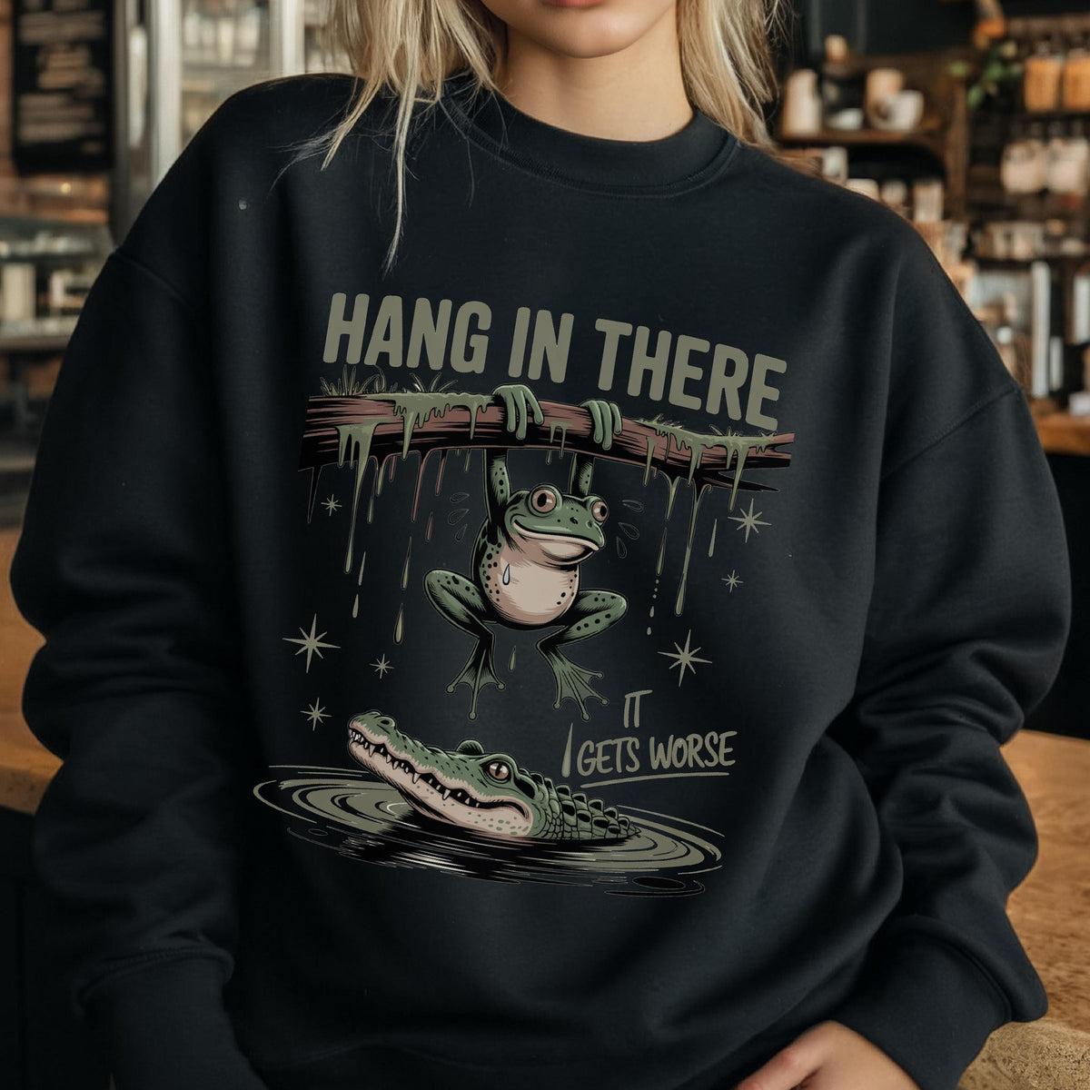 May include: Black sweatshirt with a humorous graphic. The design features a frog hanging from a branch above an alligator in water, with the text "HANG IN THERE" and "IT GETS WORSE". The frog is green and the alligator is green and gray.