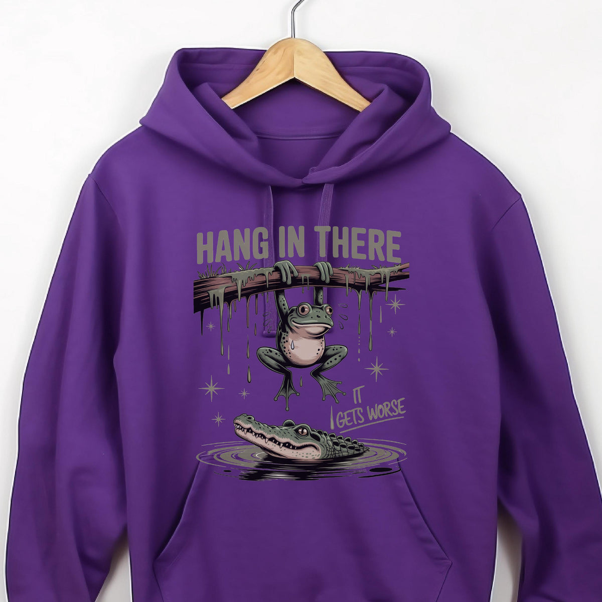 May include: A purple hoodie with a humorous graphic. The graphic features a frog hanging from a branch with the text "HANG IN THERE" above it. Below, an alligator lurks in the water, with the text "IT GETS WORSE".
