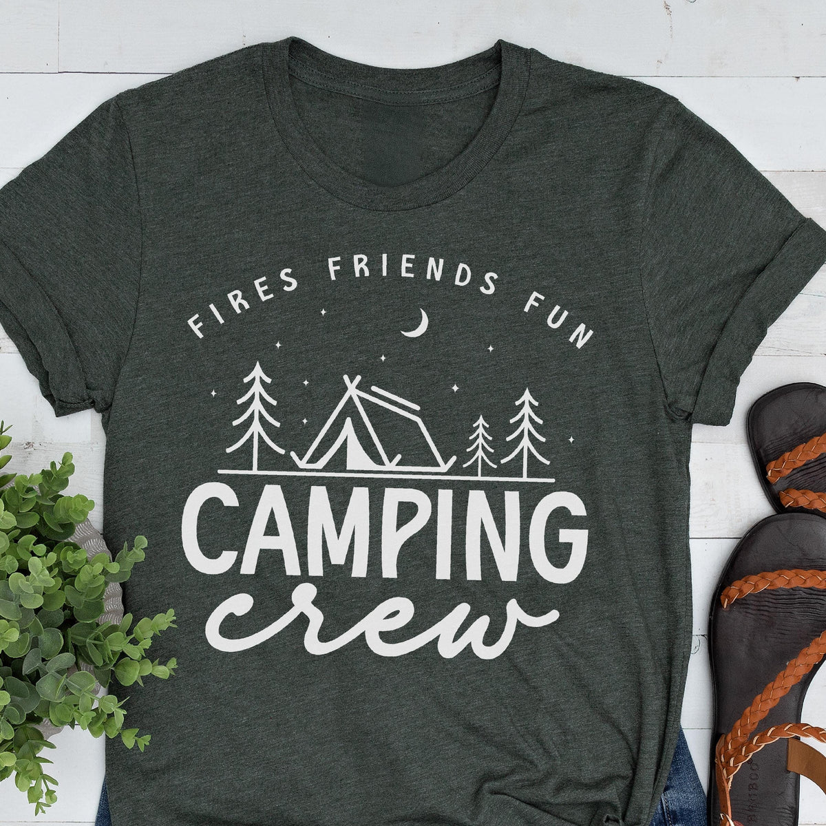 May include: Dark gray t-shirt with white text and graphics. The shirt features the words "Fires Friends Fun" above a tent and tree design, with "CAMPING crew" below. The shirt is folded, with sandals visible on the right.