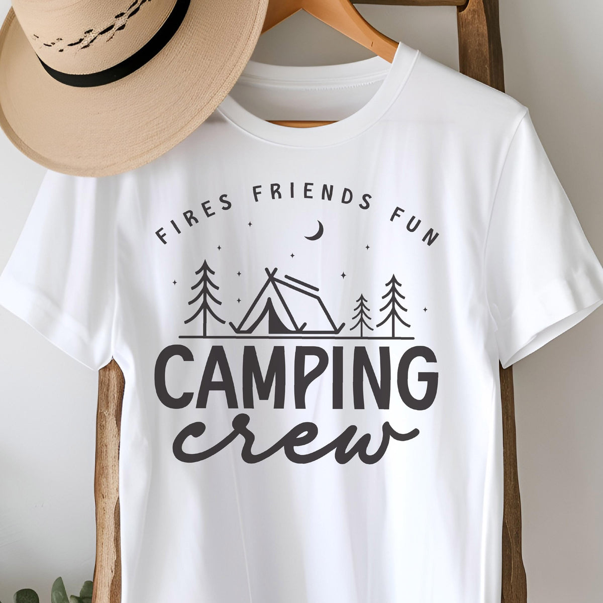 May include: White t-shirt with a black graphic design featuring a tent, trees, and the words "FIRES FRIENDS FUN" above and "CAMPING CREW" below. A tan hat hangs above the shirt.
