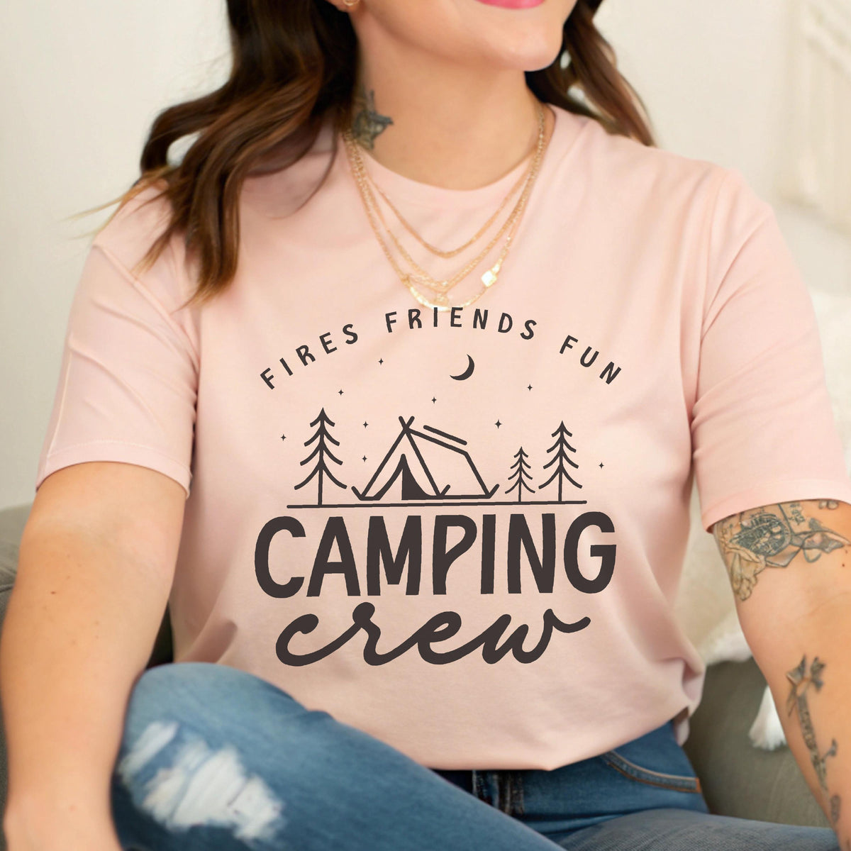May include: A light pink t-shirt with the words "FIRES FRIENDS FUN" above a camping scene with a tent and trees, and the words "CAMPING crew" below. The shirt is a soft, comfortable material.