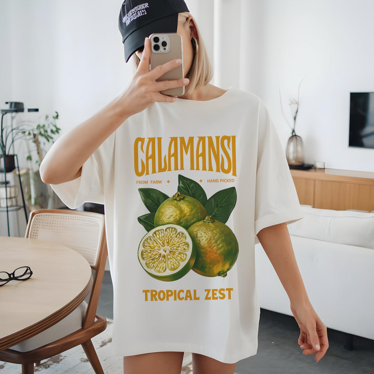 May include: Cream-colored t-shirt featuring a graphic of calamansi fruit with green leaves. The text "CALAMANSI" is above the fruit, with "FROM FARM + HAND PICKED" and "TROPICAL ZEST" below. The shirt is a casual, oversized style.