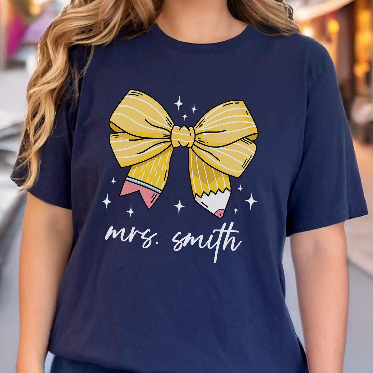 May include: Navy blue t-shirt featuring a yellow bow and pencil design with the text "mrs. smith". The bow is detailed with yellow lines and the pencils have pink and white accents. White star graphics surround the bow.