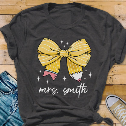 May include: Dark gray t-shirt featuring a yellow bow design made of pencils. The pencils have pink erasers and white tips. The text "mrs. smith" is written in a cursive font below the bow. White star accents are scattered around the bow.
