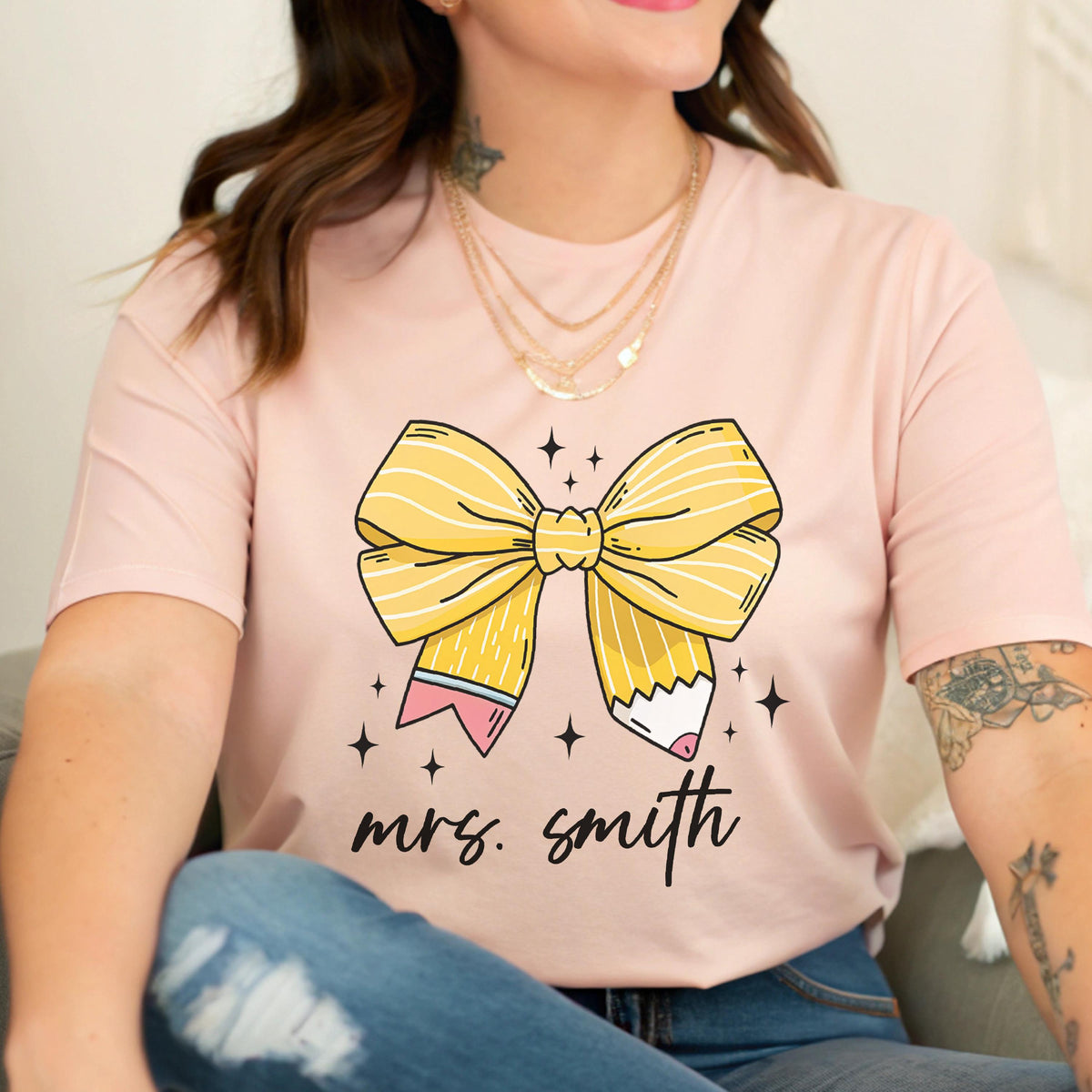 May include: A light pink t-shirt featuring a large yellow pencil bow design with the text "mrs. smith" in black script. The bow has black star accents. The shirt is a crew neck style.