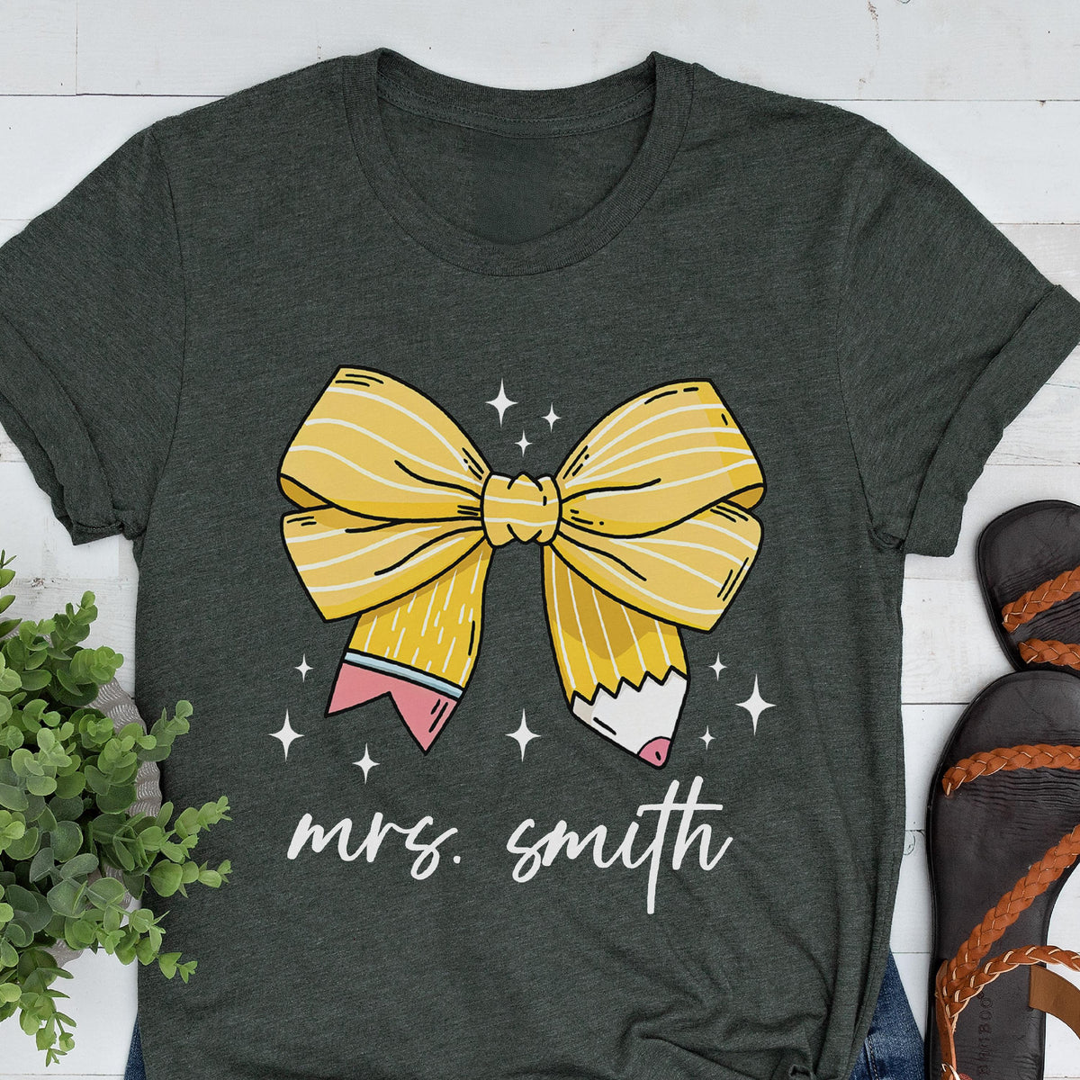 May include: Dark gray t-shirt featuring a large yellow bow design with pencil details and the text "mrs. smith" in white script. The bow has white stripes and is surrounded by small white stars. The shirt has rolled-up sleeves.