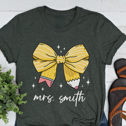 May include: Dark gray t-shirt featuring a large yellow bow design with pencil details and the text "mrs. smith" in white script. The bow has white stripes and is surrounded by small white stars. The shirt has rolled-up sleeves.