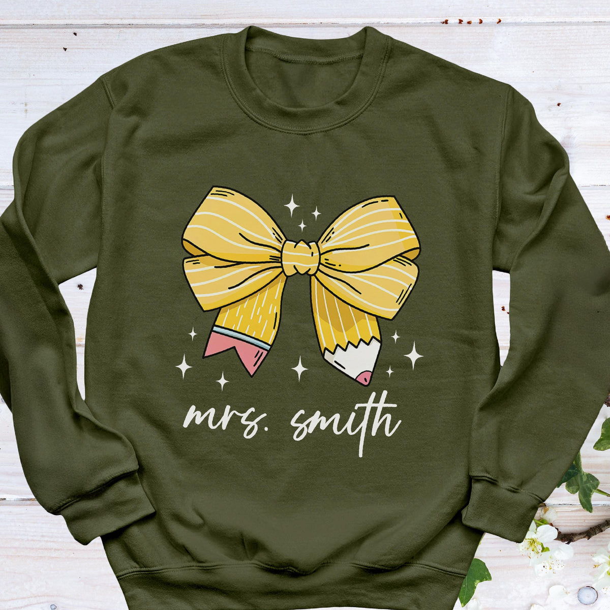 May include: Olive green sweatshirt featuring a yellow bow design made of pencils. The bow is centered on the front, with the text "mrs. smith" in a cursive font below it. White star accents are scattered around the bow.