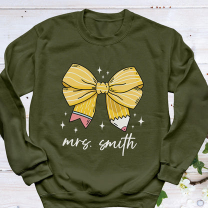 May include: Olive green sweatshirt featuring a yellow bow design made of pencils. The bow is centered on the front, with the text "mrs. smith" in a cursive font below it. White star accents are scattered around the bow.