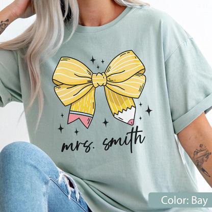 May include: A light green t-shirt with a large yellow bow and pencil design. The bow is striped and the pencils have pink erasers. The words "mrs. smith" are written in black script below the bow. The shirt is a solid color.