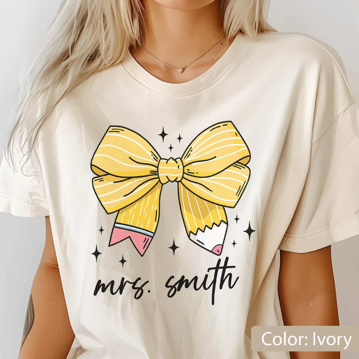 May include: Ivory-colored t-shirt featuring a large yellow bow designed as a pencil. The bow has white stripes and is accented with a pink and blue pencil tip. The words "mrs. smith" are written in black script below the bow. Small black star graphics are scattered around the bow.