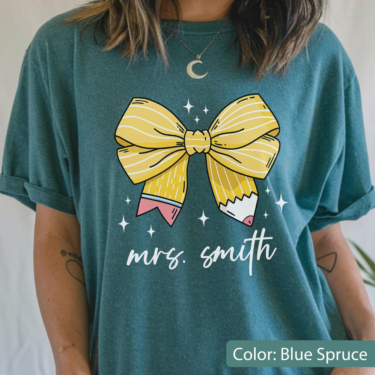 May include: Blue spruce t-shirt featuring a large yellow bow with pencil details and the text "mrs. smith" in white script. The bow design includes pink and white accents. The shirt is a solid color.