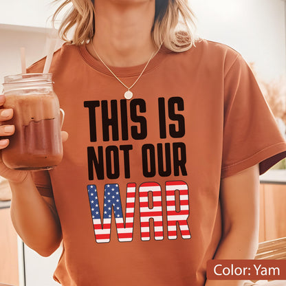 May include: A yam-colored t-shirt with the text "THIS IS NOT OUR WAR" in black block letters. The word "WAR" is designed with a red and white striped pattern and a blue star pattern.