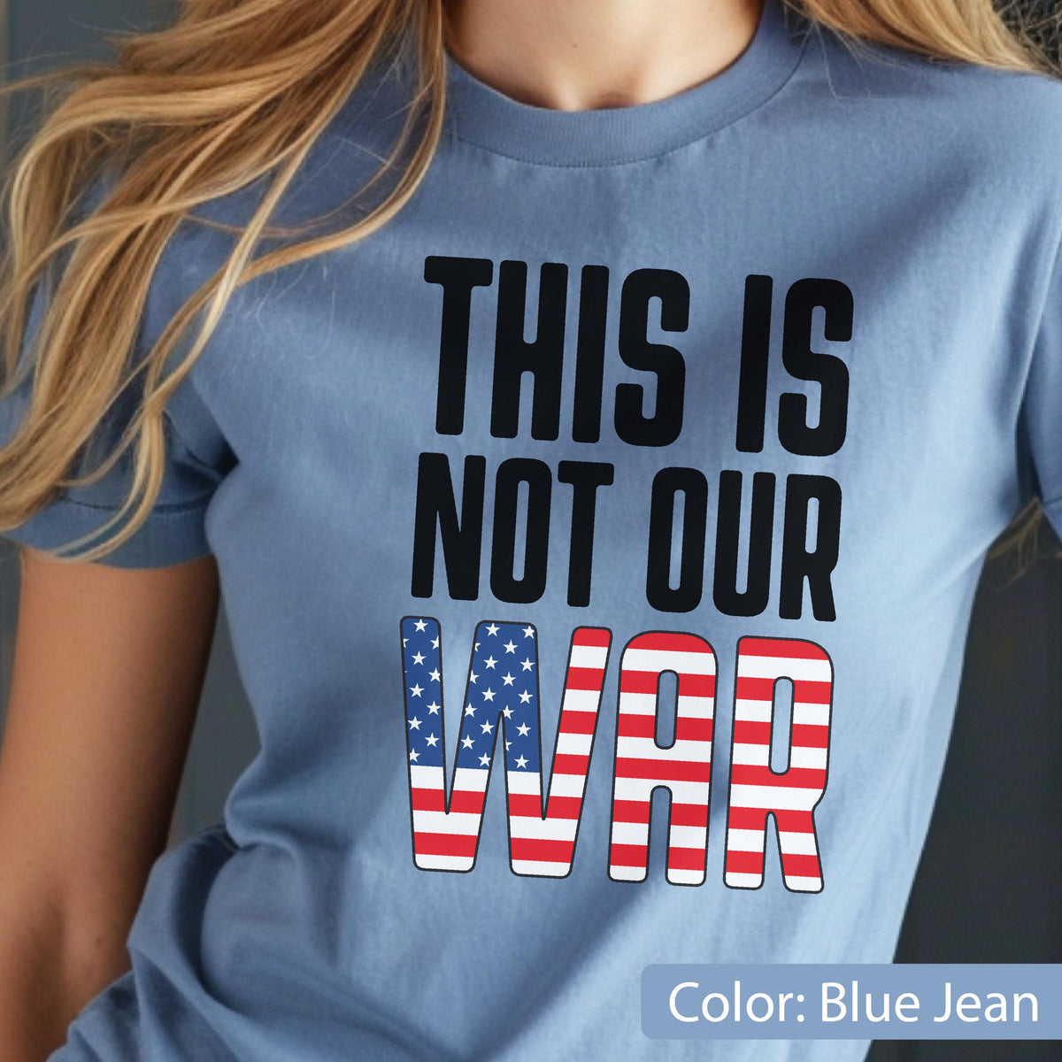 May include: Blue jean colored t-shirt with the text "THIS IS NOT OUR WAR" in black block letters. The word "WAR" is designed with the American flag pattern. The shirt is a crew neck style.