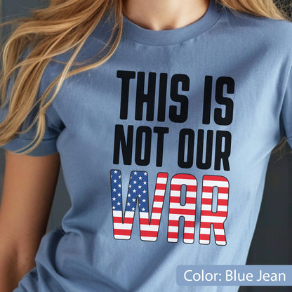 May include: Blue jean colored t-shirt with the text "THIS IS NOT OUR WAR" in black block letters. The word "WAR" is designed with the American flag pattern. The shirt is a crew neck style.