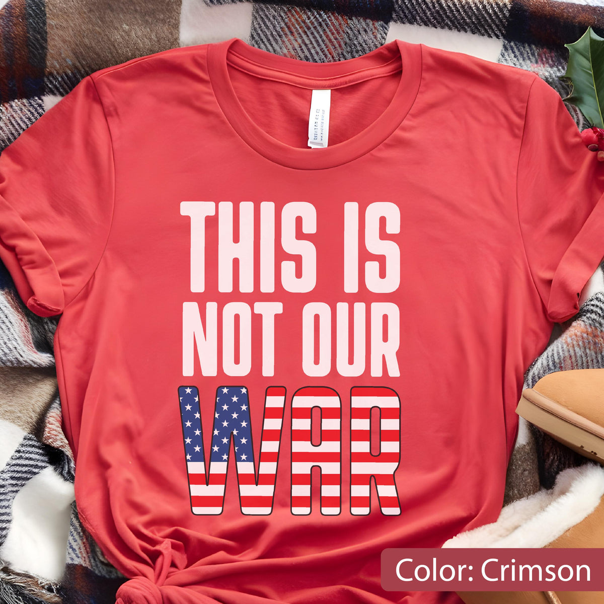 May include: Crimson-colored t-shirt with the text "THIS IS NOT OUR WAR." The word "WAR" is designed with a pattern of red and white stripes and blue stars, resembling the American flag. The shirt is folded on a plaid blanket.