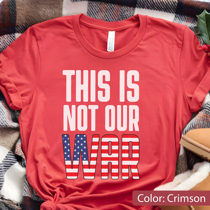 May include: Crimson-colored t-shirt with the text "THIS IS NOT OUR WAR." The word "WAR" is designed with a pattern of red and white stripes and blue stars, resembling the American flag. The shirt is folded on a plaid blanket.