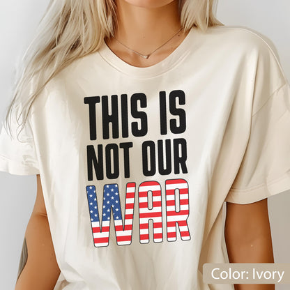 May include: Ivory-colored t-shirt with the text "THIS IS NOT OUR WAR" in black block letters. The word "WAR" is designed with the American flag's stars and stripes. The shirt is a casual, everyday wear item.