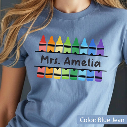 May include: Blue jean colored t-shirt with a crayon design. The design features a row of colorful crayons above and below the text "Mrs. Amelia". The shirt is a short-sleeved crew neck.