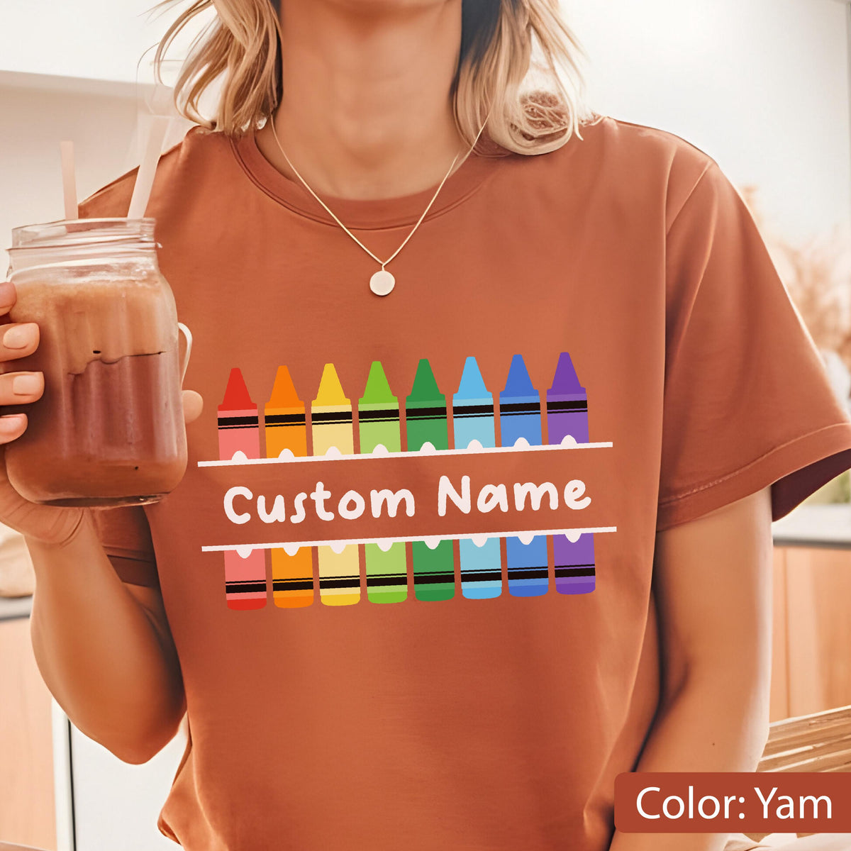 May include: A yam-colored t-shirt featuring a colorful crayon design. The design includes a row of crayons above and below the text "Custom Name". The shirt is made of a soft material.