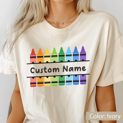 May include: Ivory-colored t-shirt featuring a colorful crayon design. Above the crayons is a horizontal banner with the text "Custom Name". The crayons are arranged in a rainbow pattern. The shirt is a neutral color.