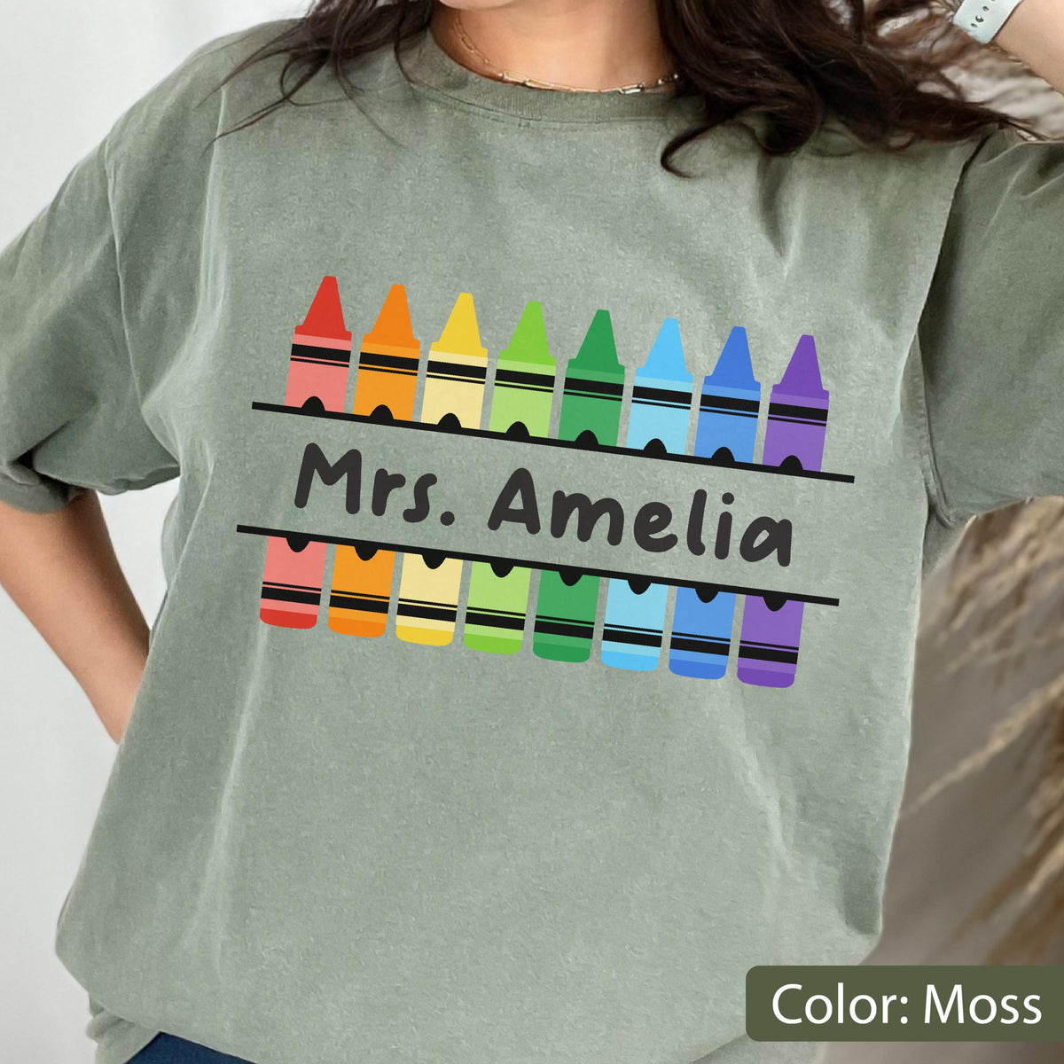 May include: Moss green t-shirt featuring a colorful crayon design above and below the text "Mrs. Amelia". The crayons are in a rainbow of colors. The shirt is a comfortable, casual style.