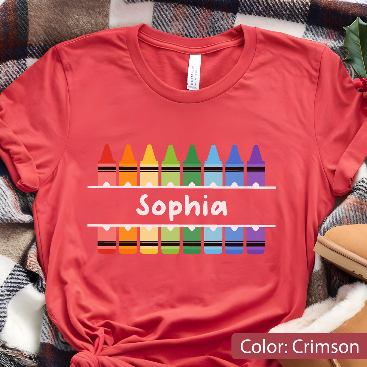 May include: Crimson red t-shirt with a colorful crayon design. The shirt features a row of crayons above and below the name "Sophia" in white. The shirt is made of a soft material.