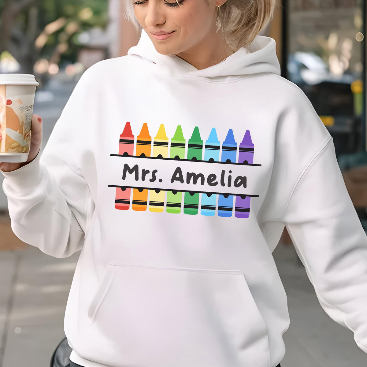 May include: White hoodie with a front pocket and a colorful crayon design. The design includes the text "Mrs. Amelia" in black, with a row of crayons above and below the text, in rainbow colors.