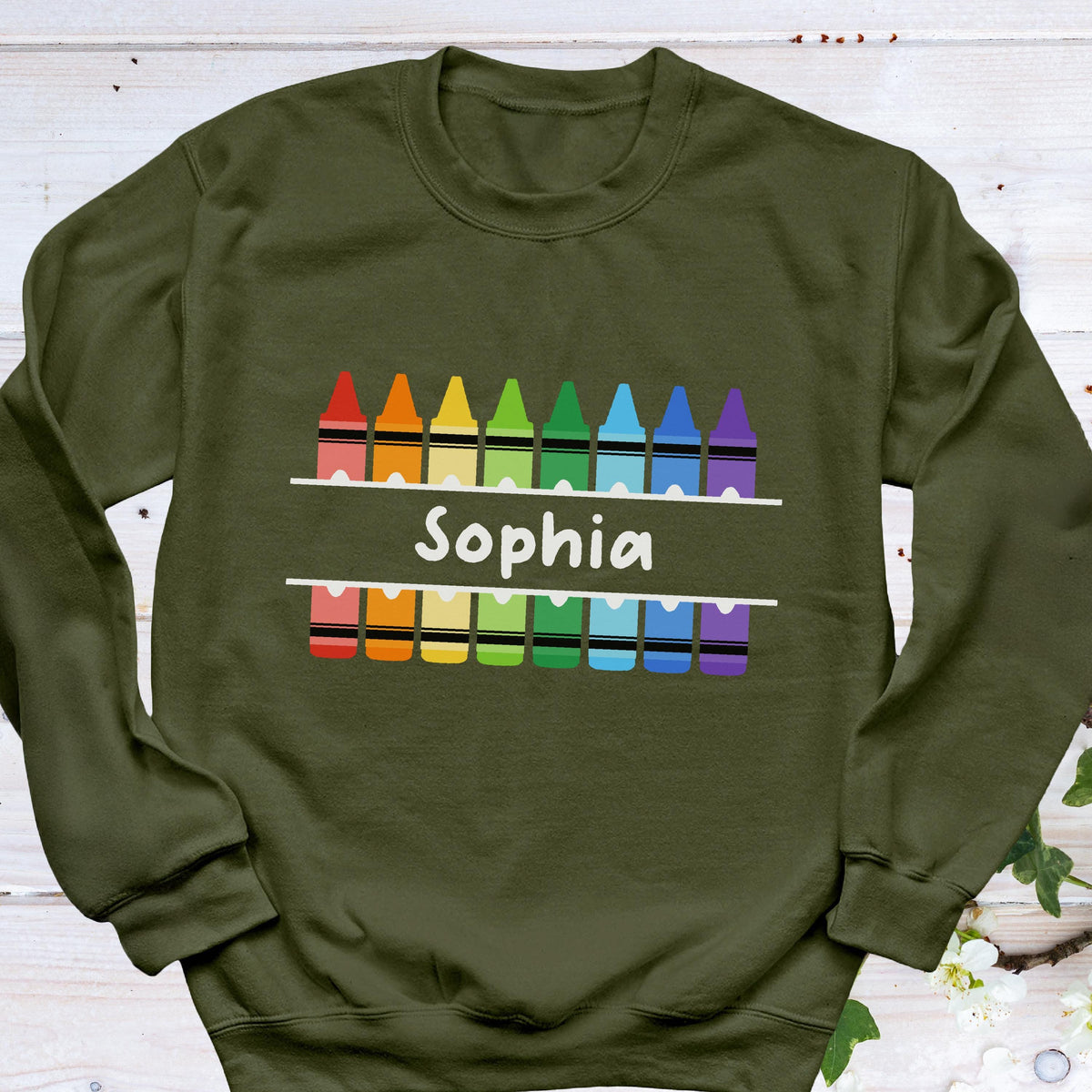 May include: Olive green sweatshirt with a colorful crayon design. The design includes a row of crayons in rainbow colors above and below the name "Sophia" in white, creating a playful and personalized look. A comfortable and stylish garment.