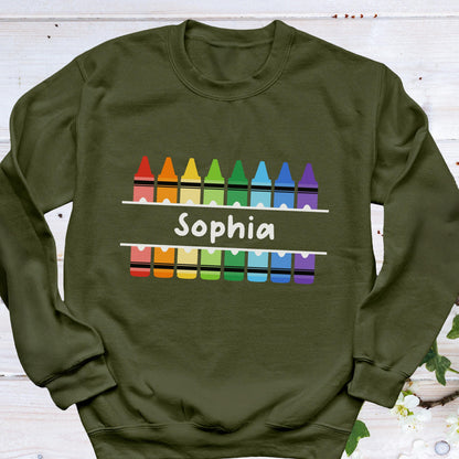 May include: Olive green sweatshirt with a colorful crayon design. The design includes a row of crayons in rainbow colors above and below the name "Sophia" in white, creating a playful and personalized look. A comfortable and stylish garment.