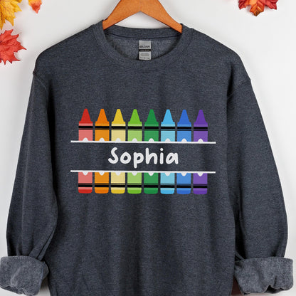 May include: A dark gray sweatshirt featuring a colorful crayon design with the name "Sophia" in white. The crayons are arranged in a rainbow pattern above and below the name, creating a vibrant and playful look.