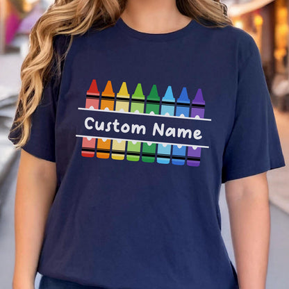 May include: Navy blue t-shirt featuring a colorful crayon design. The design includes a row of crayons above and below the text "Custom Name" in white. The crayons are in a rainbow color scheme.