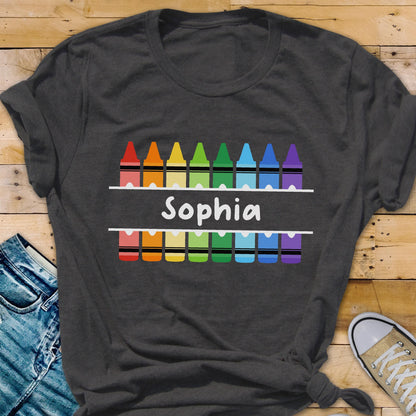May include: Dark gray t-shirt featuring a colorful crayon design with the name "Sophia" in white. The design includes a row of crayons above and below the name, creating a vibrant and playful look. The shirt is made of soft material.