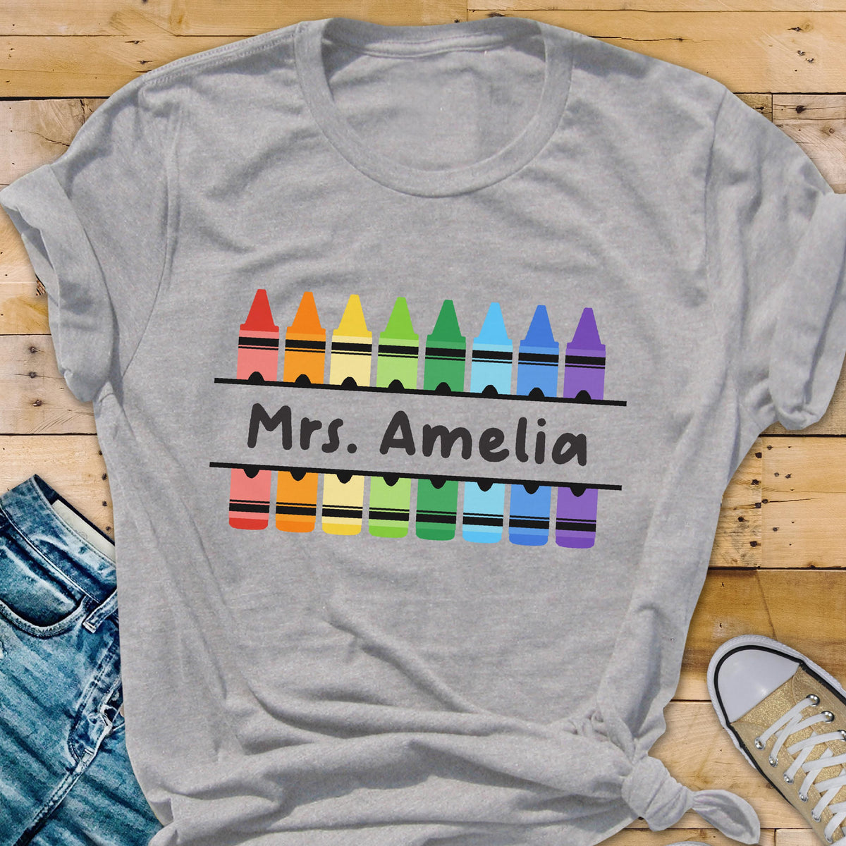 May include: A gray t-shirt featuring a colorful crayon design with the name "Mrs. Amelia" printed in black. The crayons are arranged in a row, with each crayon in a different color. The shirt is made of a soft material.