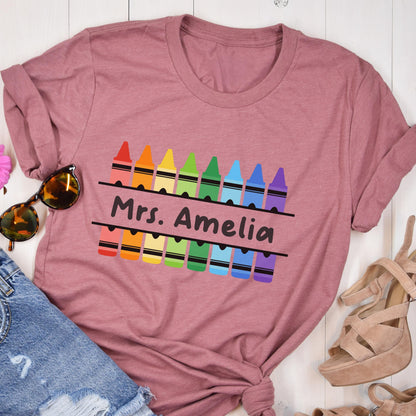 May include: A dusty rose t-shirt featuring a row of colorful crayons above and below the text "Mrs. Amelia" in black. The crayons are in a rainbow of colors. The shirt is folded and styled with sunglasses and denim shorts.