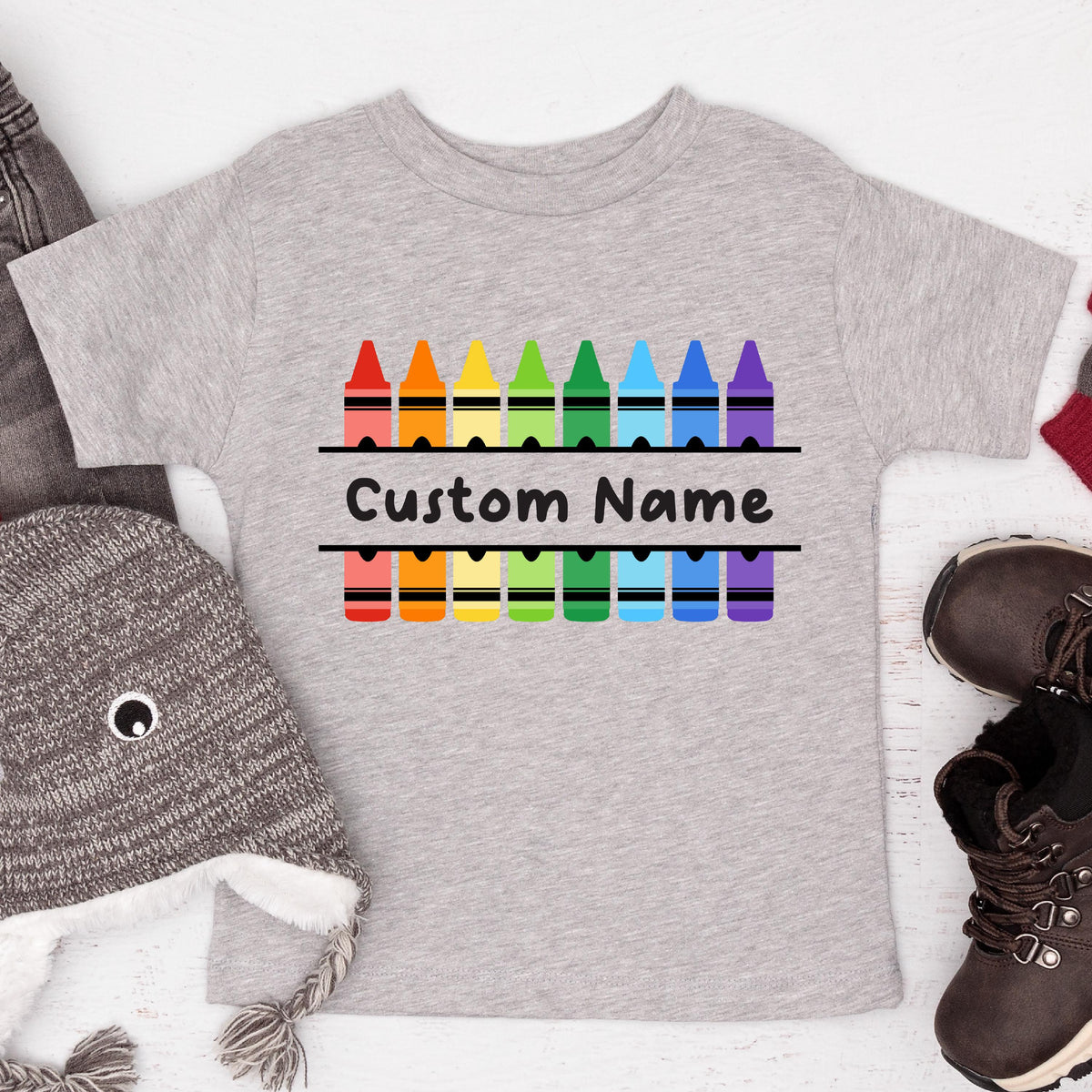 May include: A heather gray short-sleeved t-shirt featuring a graphic of two rows of colorful crayons in red, orange, yellow, green, blue, and purple. Between the rows of crayons, the text Custom Name is printed in a bold black font.