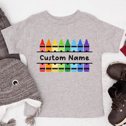 May include: A heather gray short-sleeved t-shirt featuring a graphic of two rows of colorful crayons in red, orange, yellow, green, blue, and purple. Between the rows of crayons, the text Custom Name is printed in a bold black font.