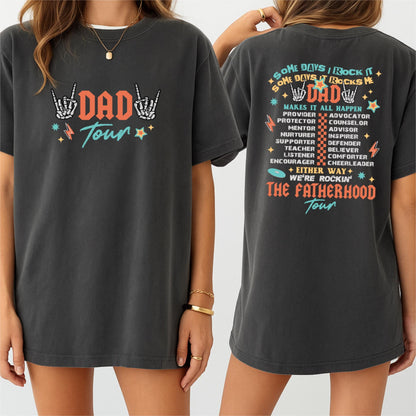 May include: Dark gray t-shirt with the text "DAD Tour" in orange and teal, featuring skeleton hand gestures and stars. The back of the shirt has the text "SOME DAYS I ROCK IT, DAD, MAKES IT ALL HAPPEN" and a list of fatherly roles.