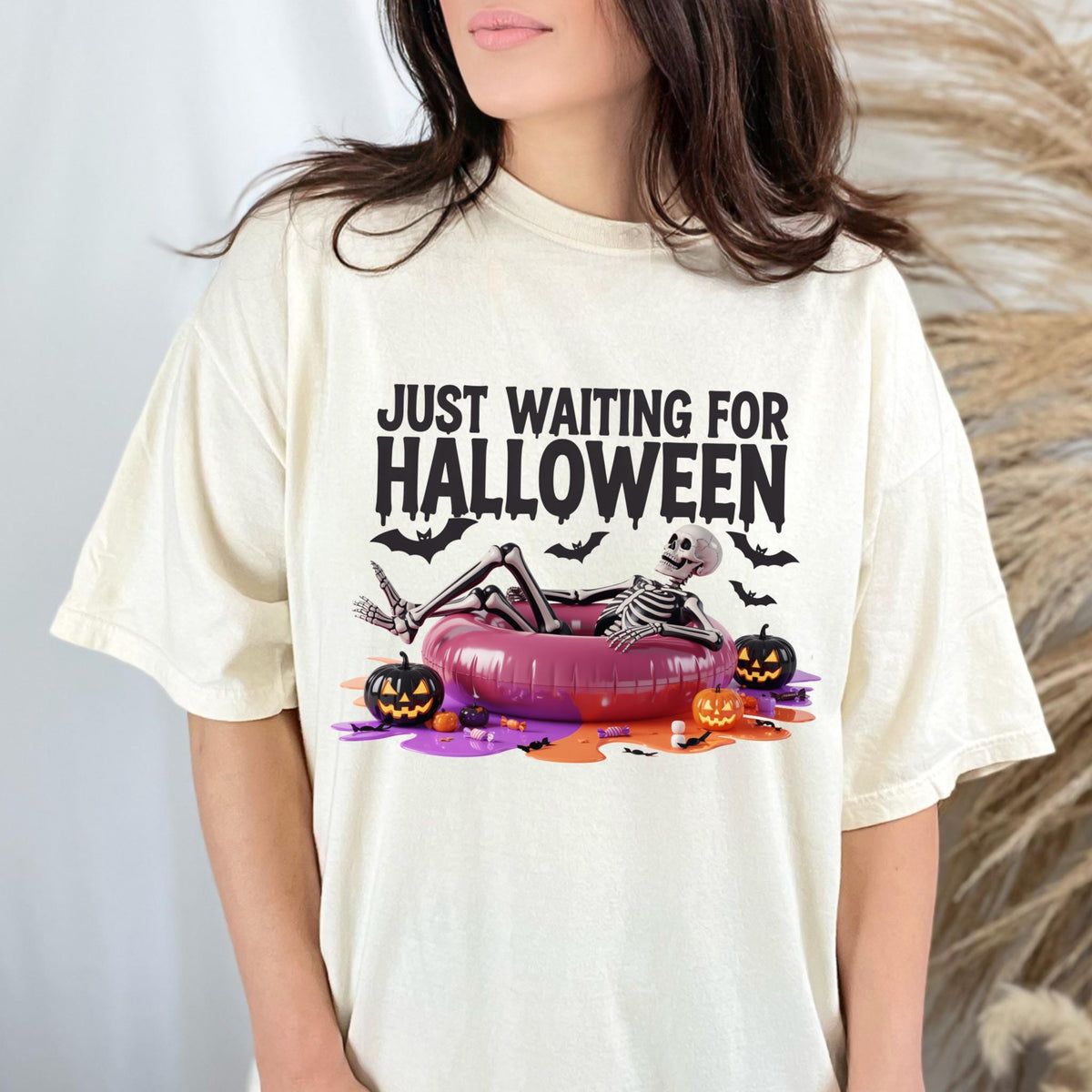 May include: Cream-colored t-shirt with the text "JUST WAITING FOR HALLOWEEN". The graphic shows a skeleton relaxing in a pink inflatable ring, surrounded by bats and jack-o'-lanterns. A fun, festive design for Halloween.