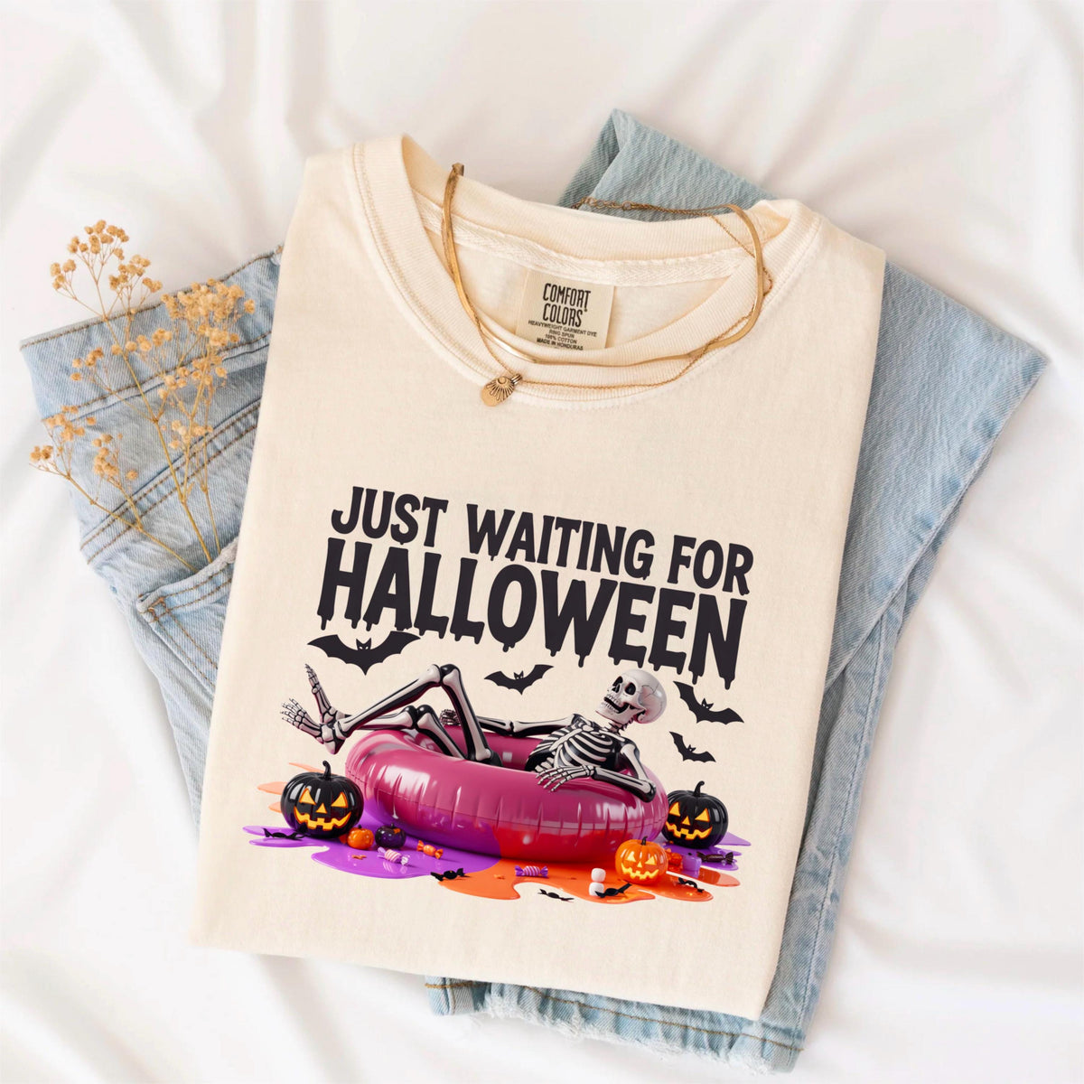 May include: Cream-colored t-shirt with the text "JUST WAITING FOR HALLOWEEN" and a skeleton relaxing in a pink inflatable ring. The design includes bats and jack-o'-lanterns, perfect for Halloween. The shirt is folded on top of blue jeans.