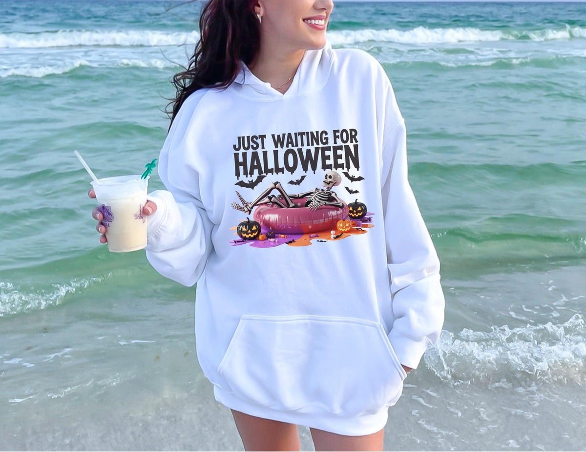 May include: White hooded sweatshirt with a front pocket and the text "JUST WAITING FOR HALLOWEEN". The graphic features a skeleton relaxing on a pool float with pumpkins and bats. The person is holding a drink.