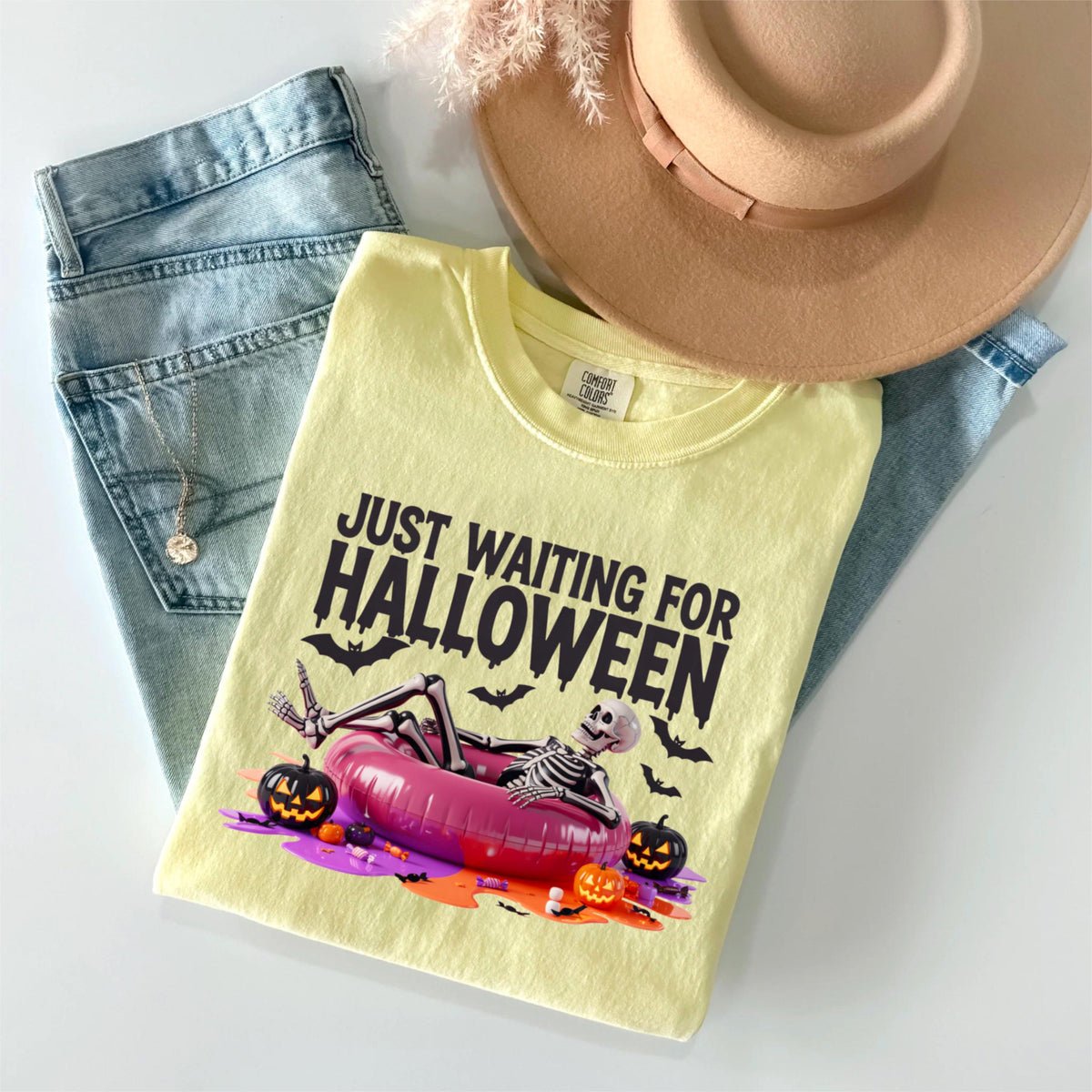 May include: A light yellow Comfort Colors t-shirt with the text "JUST WAITING FOR HALLOWEEN" and a skeleton relaxing in a pink inflatable ring. The shirt is paired with light blue jeans and a tan hat. Two black jack-o'-lanterns and bats are also featured.
