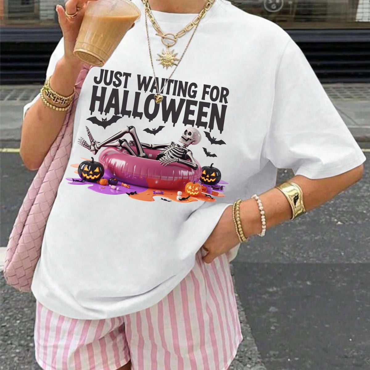 May include: White t-shirt with the text "JUST WAITING FOR HALLOWEEN". The graphic features a skeleton relaxing in a pink inflatable ring, surrounded by pumpkins and bats. The shirt is paired with pink and white striped shorts.